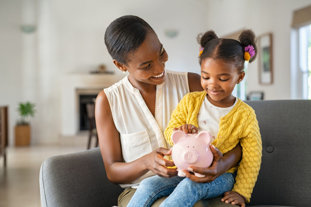 Smiling mature african american mother helping daughter sitting on lap putting money in piggy bank.