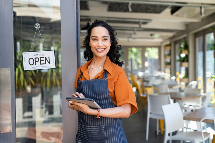 Happy young latin woman owner in apron standing at coffee shop entrance, leaning while waiting clients. Successful cafe business owner looking away with copy space.